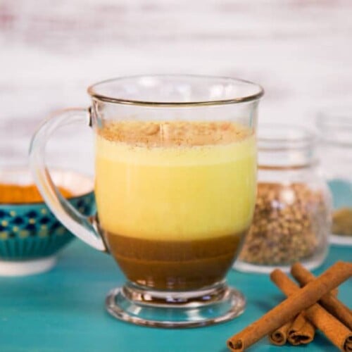 A glass mug containing a layered turmeric chai latte, with vibrant yellow on top blending into rich brown below, accompanied by a bowl on the left and spice jars to the right.