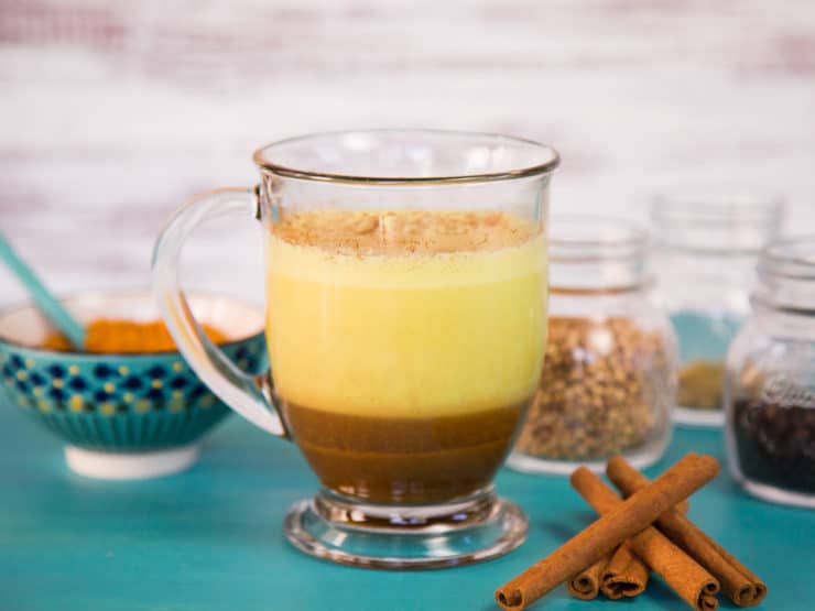 A glass mug containing a layered turmeric chai latte, with vibrant yellow on top blending into rich brown below, accompanied by a bowl on the left and spice jars to the right.