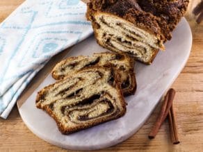 Vertical shot - loaf of tender cinnamon babka with two slices on white cutting board, sticks of cinnamon and cloth napkin alongside.