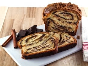 Frontal shot of sliced babka with swirling chocolate filling, chocolate chunks and cinnamon sticks, and cloth napkin on white cutting board with wood background.