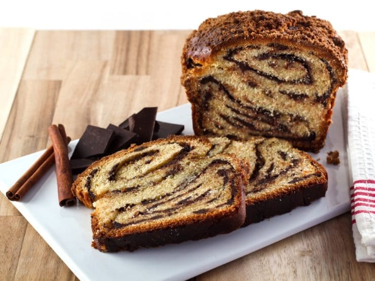 Frontal shot of sliced babka with swirling chocolate filling, chocolate chunks and cinnamon sticks, and cloth napkin on white cutting board with wood background.