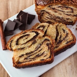 Horizontal shot of sliced babka with swirling chocolate filling, chocolate chunks on white cutting board with wood background.