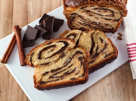 Horizontal shot of sliced babka with swirling chocolate filling, chocolate chunks on white cutting board with wood background.