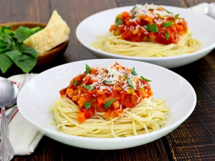 Horizontal crop - plate of spaghetti pasta with pomodoro sauce topped with basil and parmesan, cloth napkin with utensils beside it, another plate of pasta, parmesan block and fresh basil in background.