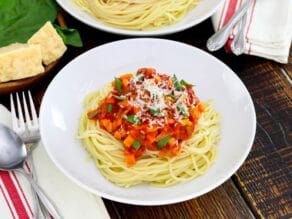 Overhead shot - close up, white plate of spaghetti pasta with cooked pomodoro tomato sauce topped with fresh basil and parmesan, cloth napkin with utensils beside it, another plate of pasta, parmesan block and fresh basil in background.
