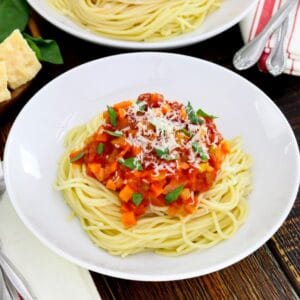 Overhead shot - close up, white plate of spaghetti pasta with cooked pomodoro tomato sauce topped with fresh basil and parmesan, cloth napkin with utensils beside it, another plate of pasta, parmesan block and fresh basil in background.