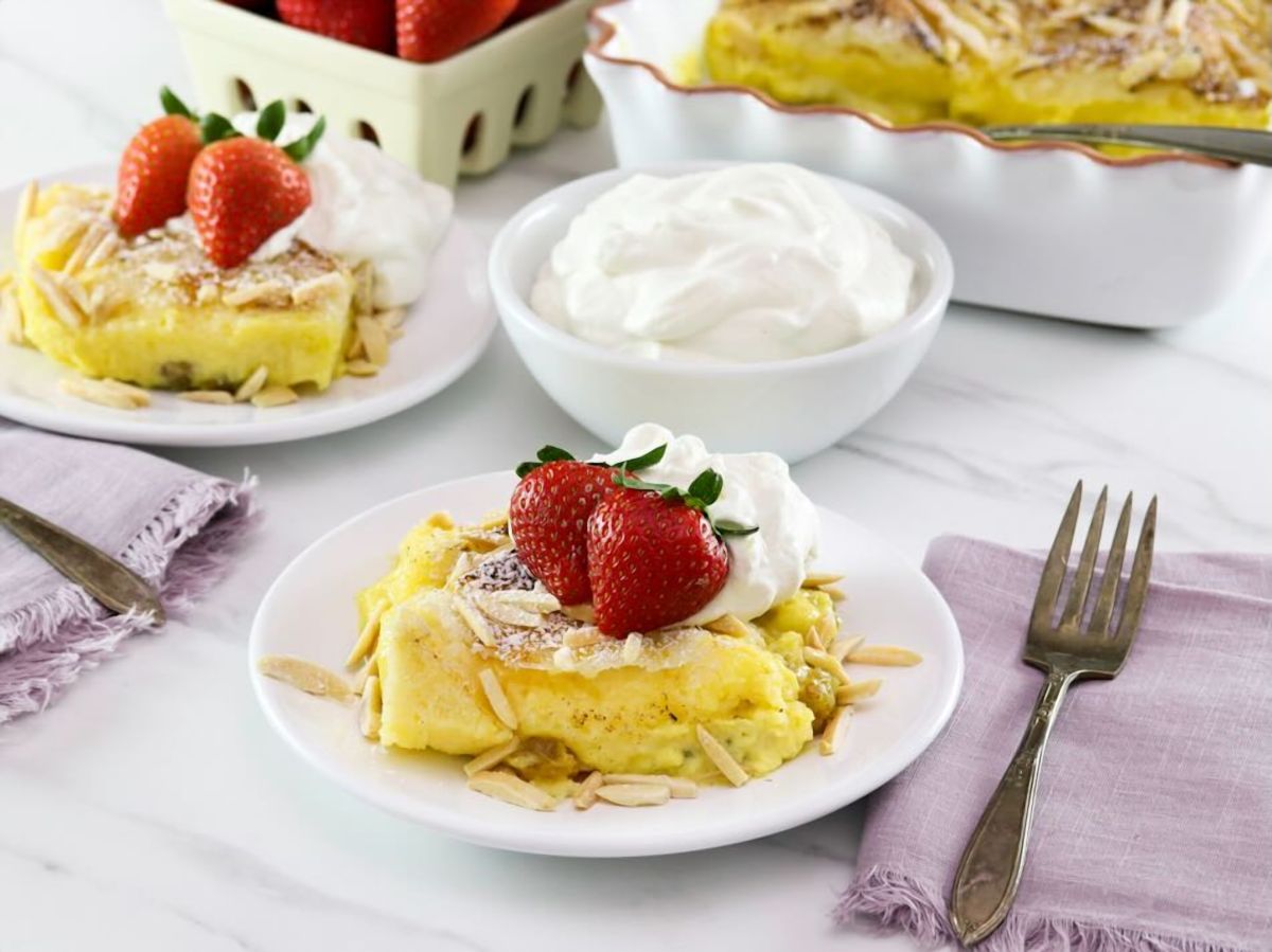 Horizontal shot - a delicious slice of Princess Diana's favorite recipe - bread and butter pudding topped with fresh strawberries and whipped cream. Dish of whipped cream. additional serving, and berries in background.