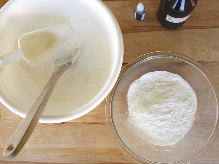 White mixing bowl with powder, scoop and wooden spoon, glass mixing bowl with dry ingredients on wooden cutting board.