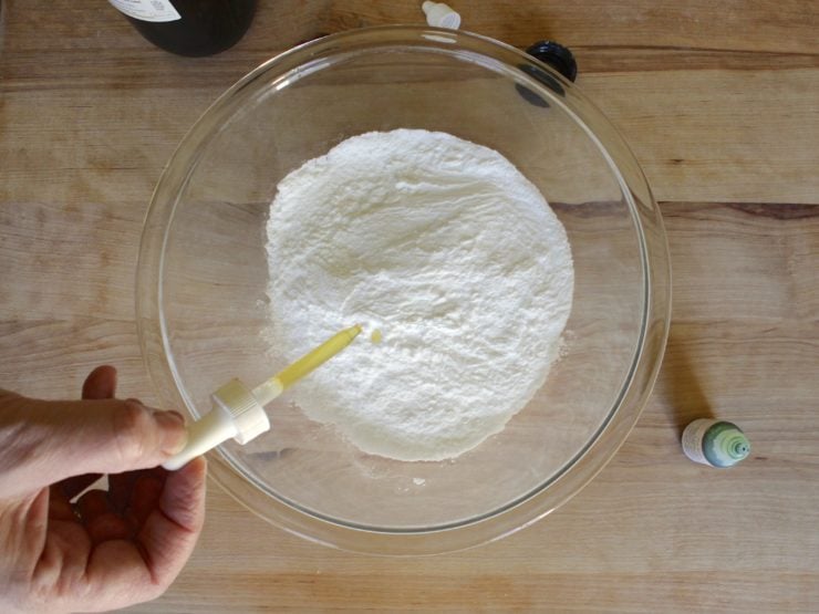 Adding dropper of scent to dry ingredients in glass mixing bowl.