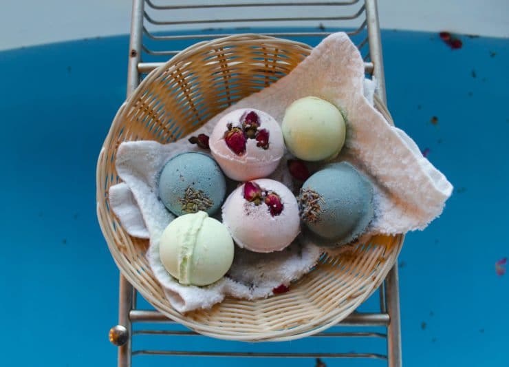 Completed bath fizzies in a variety of colors - pink, blue and green, dotted with botanicals, in basket over blue water in bathtub.