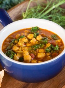 A bowl of Chickpea Kale and Fire Roasted Tomato Soup with vegetables and herbs on a cutting board
