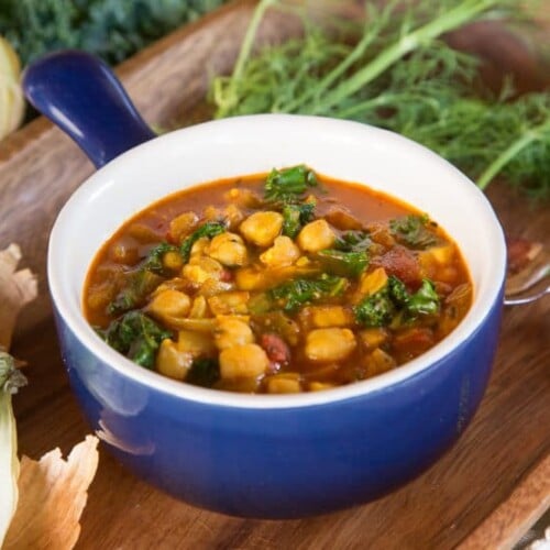 A bowl of Chickpea Kale and Fire Roasted Tomato Soup with vegetables and herbs on a cutting board