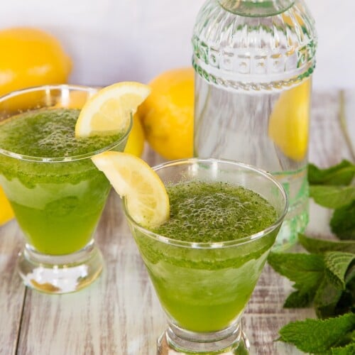 Close-up of two glasses of spiked Limonana served with a slice of lemon garnish surrounded with lemons, a tall fancy glass, and mint leaves