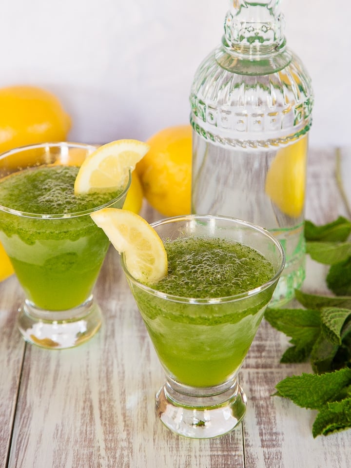 Close-up of two glasses of spiked Limonana served with a slice of lemon garnish surrounded with lemons, a tall fancy glass, and mint leaves