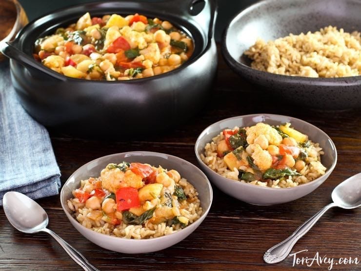 Two white bowls of curried vegetable stew next to two spoons on top of a wooden table. In the background is a large pot of stew and a bowl of rice.