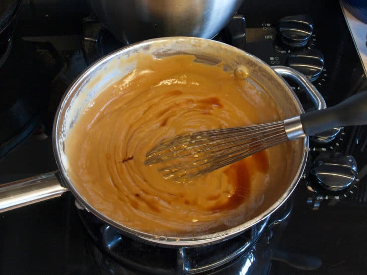 A saucepan on a stove contains smooth, creamy caramel sauce with vanilla being whisked
