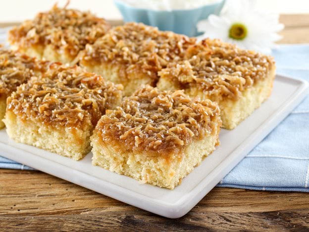 Square slices of yellow cake topped with caramelized sugar and shredded coconut on a white serving plate atop a blue placemat on a wooden background.