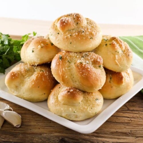 Stocked Garlic Knots topped with cheese and parsley served on a white plate surrounded with parsley and garlic cloves