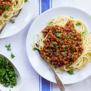 Plate of pasta topped with a lentil sauce and garnished with chopped green herbs on top of a blue and white tablecloth.