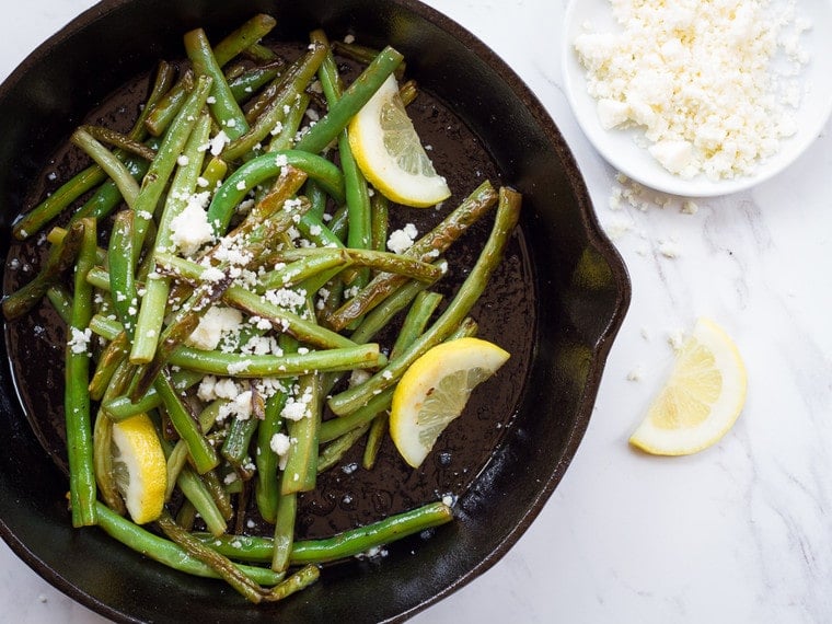 Seared green beans and cotija cheese with lemon wedges in a black cast iron skillet on a white marble background.