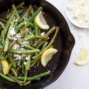 Seared green beans and cotija cheese with lemon wedges in a black cast iron skillet on a white marble background.