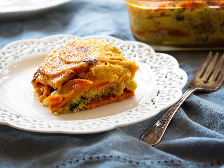 A small serving of yellow and orange Sweet Potato, Spinach and Quinoa Gratin on a white plate next to a silver fork atop a blue placemat.