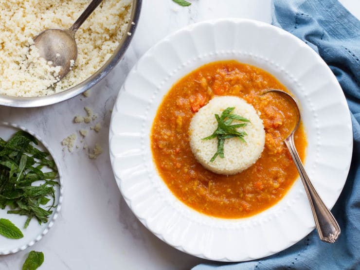 Red soup with a scoop of white rice and a green herb garnish in a white bowl next to a blue towel on a white marble background.