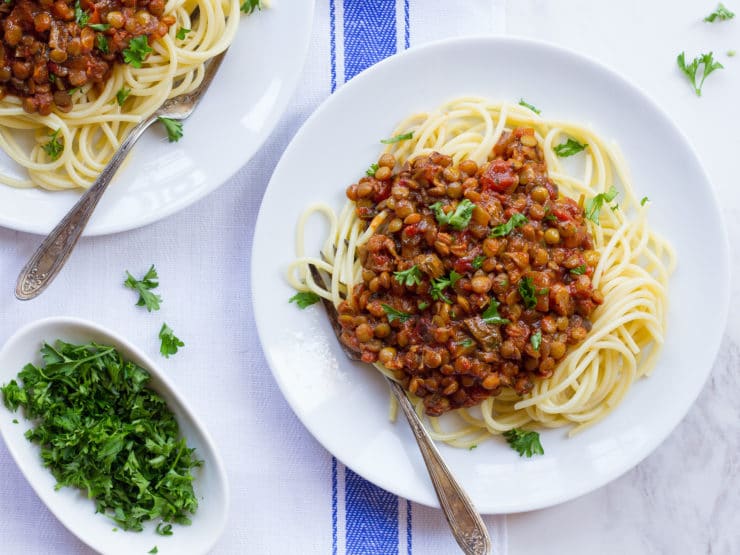 Plate of pasta topped with a lentil sauce and garnished with chopped green herbs on top of a blue and white tablecloth.