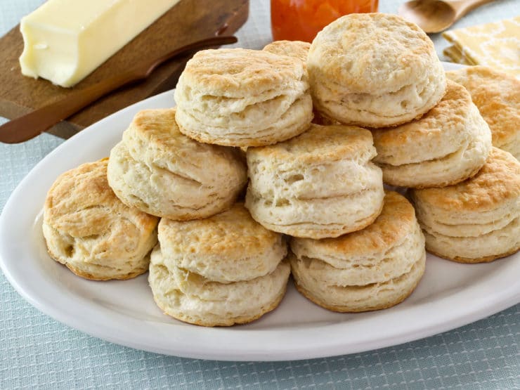 A pile of freshly baked biscuits sitting on a white plate with a stick of butter on a wooden tray in the background.