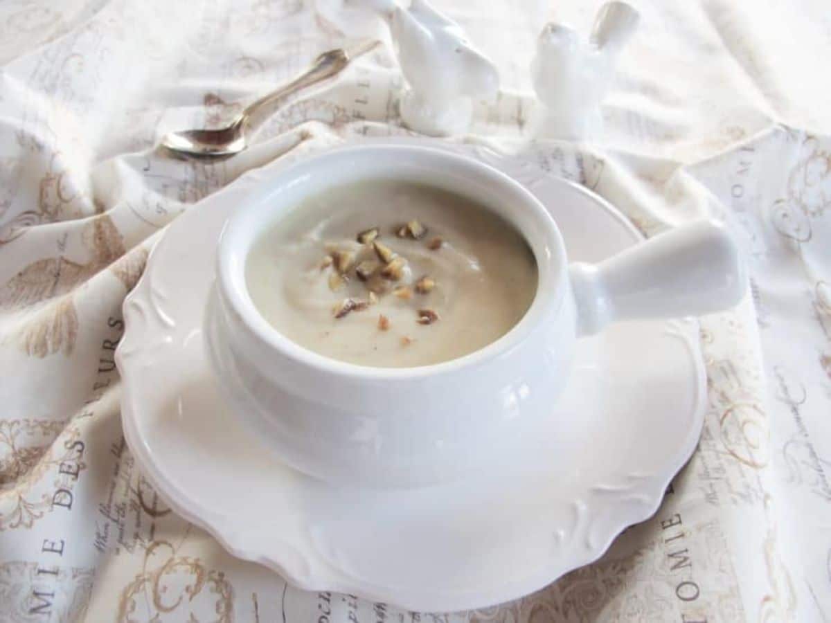 Jerusalem artichoke soup in a white bowl on a small white plate next to a silver spoon on a white cloth background.