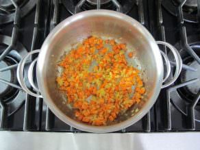 Diced carrots and onions sautéed in a stainless steel pot on the stovetop.