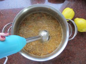 Immersion blender blending lentil soup in a stainless pot on countertop, lemon on counter.
