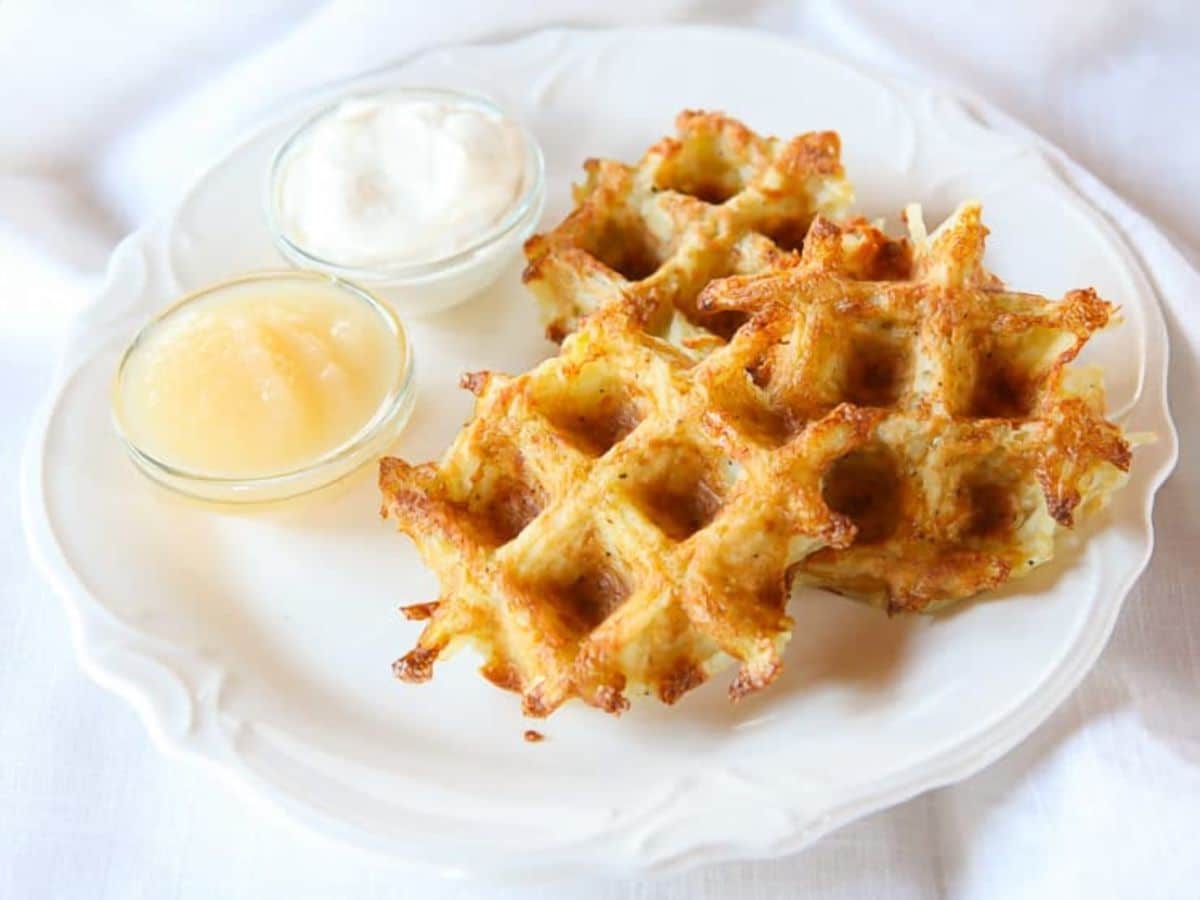 Latke waffles on a white plate with a small dish of sour cream and a small dish of apple sauce on a white cloth background.