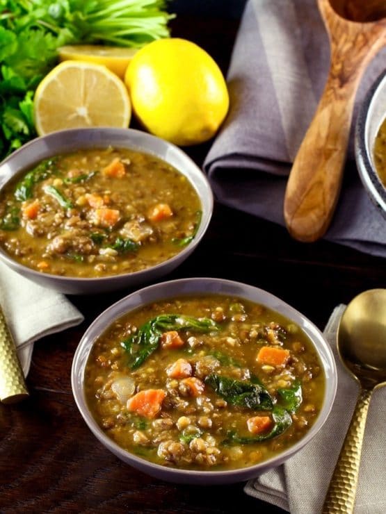 Two bowls of lentil spinach soup with spoon and napkin, lemons and parsley in background with wooden spoon.