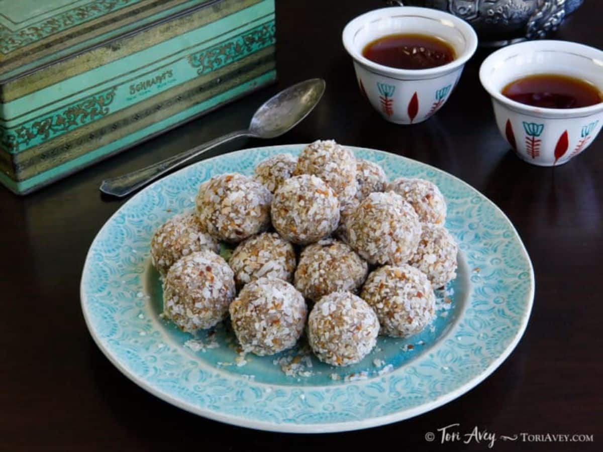 A pile of date truffles, rolled in toasted coconut, on a blue plate on top of a wooden table. In the background there are two cups of tea, a spoon, and an antique tea box.