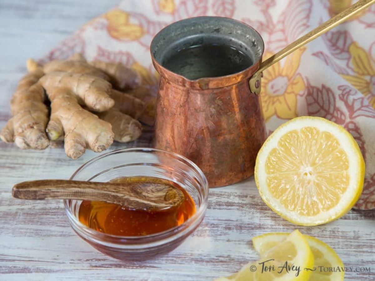 Bronze Turkish coffee maker next to fresh ginger, a fresh sliced lemon, and a small glass bowl of honey on top of a white wooden background.