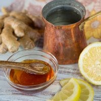 Image of a brown steel mug with a small bowl of honey, accompanied by a lemon wedge on the right and a ginger beside the mug.