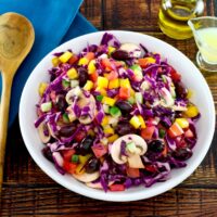 Overhead shot of bowl of colorful rainbow salad with peppers, cucumbers, mushrooms and cabbage in a white bowl with glass dish of lemon juice, carafe of olive oil and blue napkin with wooden spoon in the background.