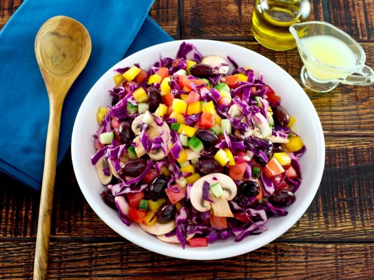 Overhead shot of bowl of colorful rainbow salad with peppers, cucumbers, mushrooms and cabbage in a white bowl with glass dish of lemon juice, carafe of olive oil and blue napkin with wooden spoon in the background.