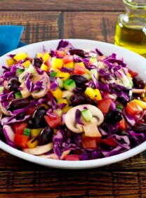 Bowl of colorful rainbow salad with peppers, cucumbers, mushrooms and cabbage in a white bowl with glass dish of lemon juice, carafe of olive oil and blue napkin with wooden spoon in the background.