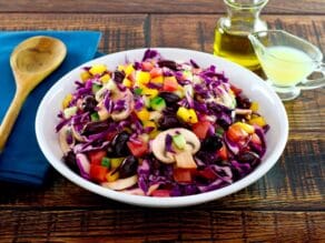 Bowl of colorful rainbow salad with peppers, cucumbers, mushrooms and cabbage in a white bowl with glass dish of lemon juice, carafe of olive oil and blue napkin with wooden spoon in the background.