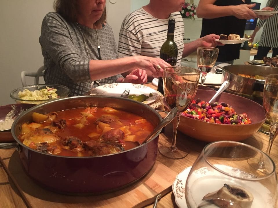 Guests enjoying a Shabbat dinner at table set with paparika pepper fish, salad, wine, and wine glasses. 