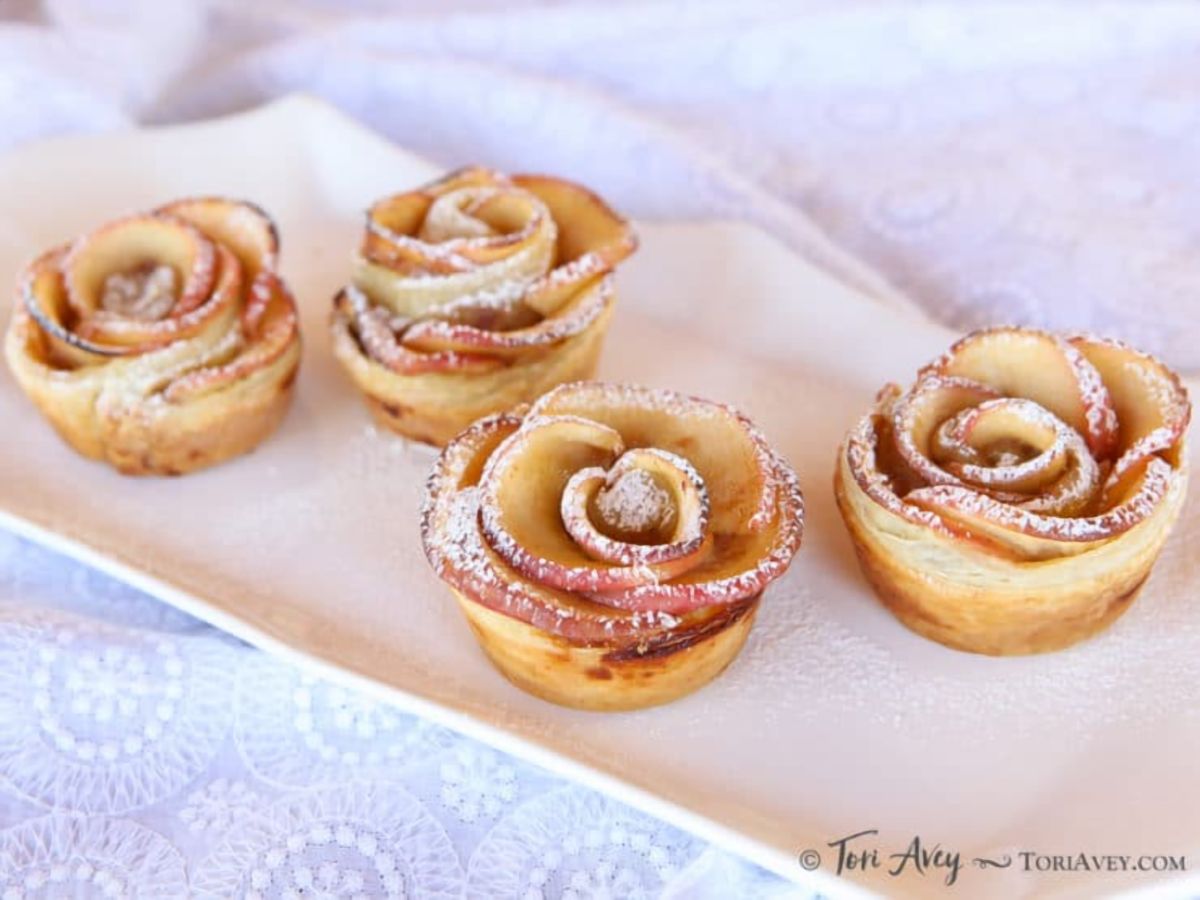 Close-up of four Apple Date Rose Tarts a vegan appetizer served on a white long fancy plate on top of a lace designed table clothe