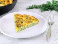 Broccoli Cheddar Frittata Pinterest Pin - slice of broccoli cheddar frittata on a small white plate with a fork, skillet of frittata, sprig of fresh dill, and a white canister in the background.