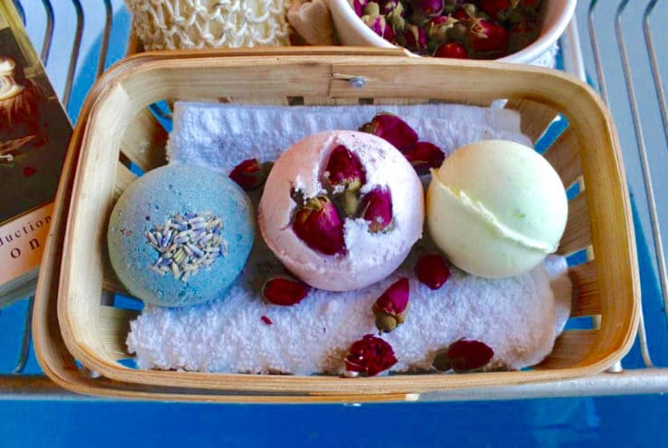 Three botanical bath fizzies resting on towel with flower petals in basket on wire shelf over blue bathwater.