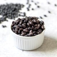 White dish of cooked black beans on marble background, dried black beans scattered behind.