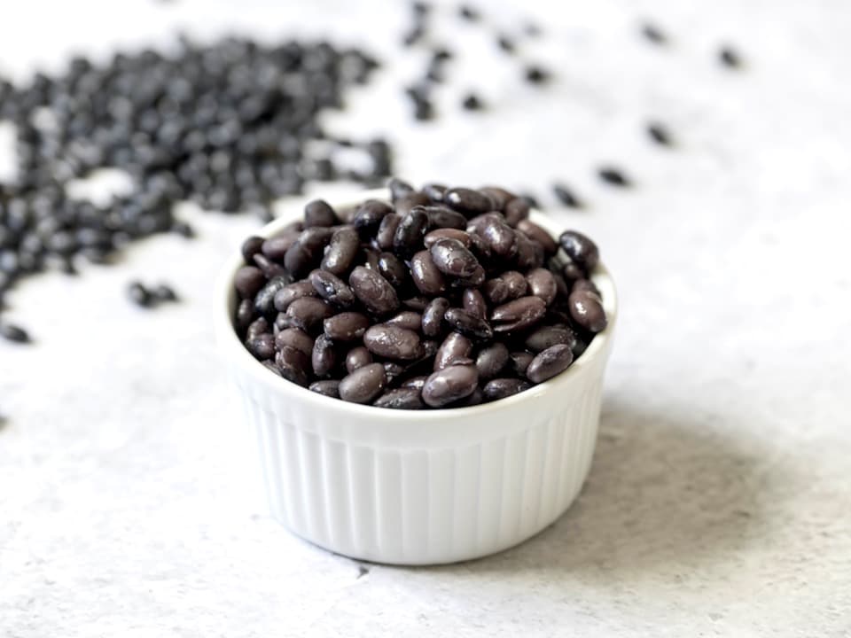 White dish of cooked black beans on marble background, dried black beans scattered behind.