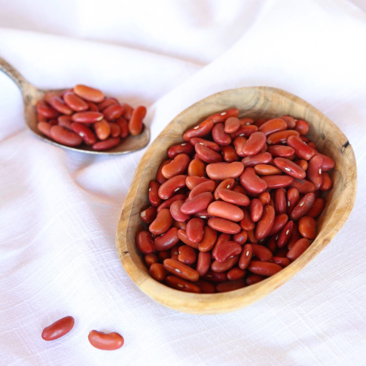 Square Crop - wooden dish of dried Kidney Beans on a white cloth background, spoon of beans in background.