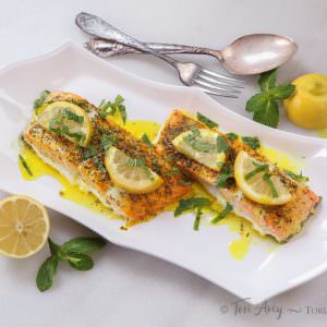 Two pieces of cooked salmon topped with lemon slices, fresh mint, and a buttery sauce on a white plate on a white background.