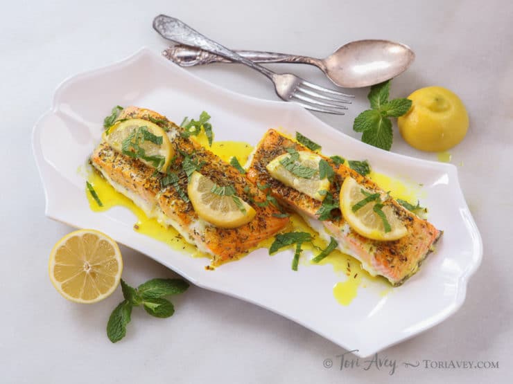 Two pieces of cooked salmon topped with lemon slices, fresh mint, and a buttery sauce on a white plate on a white background.
